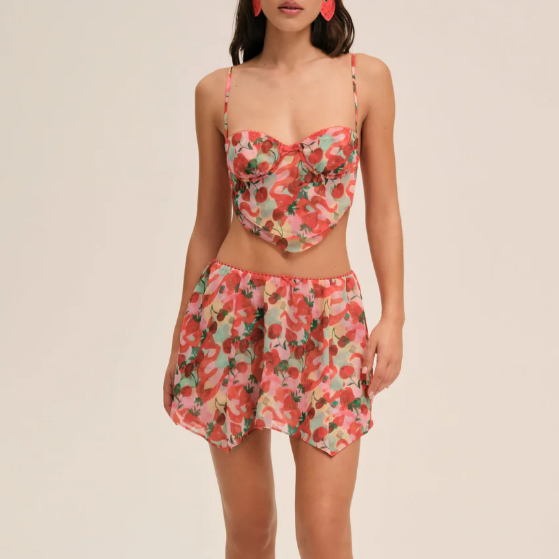 A model wearing a two-piece floral print set consisting of a sweetheart bustier crop top with spaghetti straps and a matching high-waisted mini skirt, against a solid light beige background.