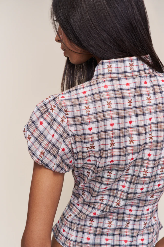 A woman shown from the back wearing a short-sleeve plaid shirt with red hearts and brown star motifs on a beige, white, and black plaid base.